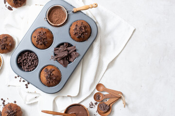 Close up top view of chocolate muffins flat lay in baking tray with slides of chocolate, chocolate chip and cocoa powder on white cutting board and white cloth with copy spacec