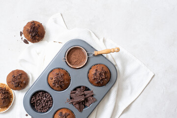 Close up top view of chocolate muffins flat lay in baking tray with slides of chocolate, chocolate chip and cocoa powder on white cutting board and white cloth with copy space