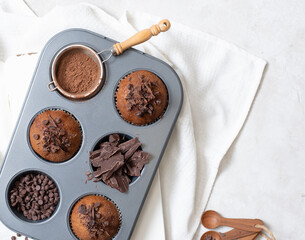 Close up top view of chocolate muffins flat lay in baking tray with slides of chocolate, chocolate chip and cocoa powder on white cutting board and white cloth
