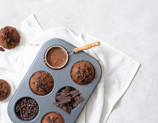 Close up top view of chocolate muffins flat lay in baking tray with slides of chocolate, chocolate chip and cocoa powder on white cutting board and white cloth with copy space