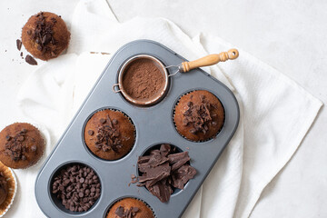 Close up top view of chocolate muffins flat lay in baking tray with slides of chocolate, chocolate chip and cocoa powder on white cutting board and white cloth