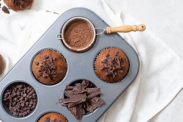 Close up top view of chocolate muffins flat lay in baking tray with slides of chocolate, chocolate chip and cocoa powder on white cutting board and white cloth
