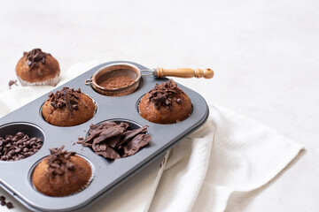 Chocolate muffins flat lay in baking tray with slides of chocolate, chocolate chip and cocoa powder on white cutting board and white cloth in soft focus