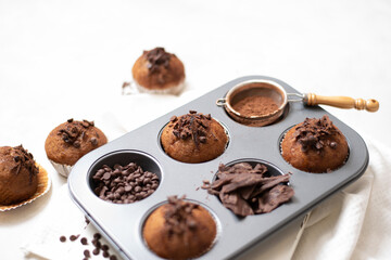 Chocolate muffins flat lay in baking tray with slides of chocolate, chocolate chip and cocoa powder on white cutting board and white cloth in soft focus