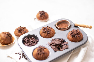 Chocolate muffins flat lay in baking tray with slides of chocolate, chocolate chip and cocoa powder on white cutting board and white cloth in soft focus