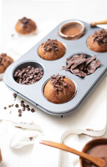 Chocolate muffins flat lay in baking tray with slides of chocolate, chocolate chip, cocoa powder and chocolate sauce on white cutting board and white cloth in soft focus