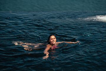 Happy woman in swimsuit swimming in infinity pool against seafront.