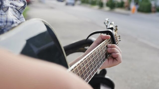 A teenage boy plays an acoustic guitar outdoors close to the road where cars pass and people pass. Teen street guitarist.