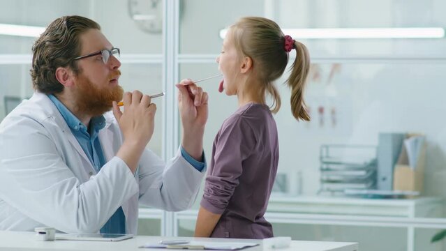Male pediatrician using tongue depressor and flashlight while giving medical checkup to little girl in clinic
