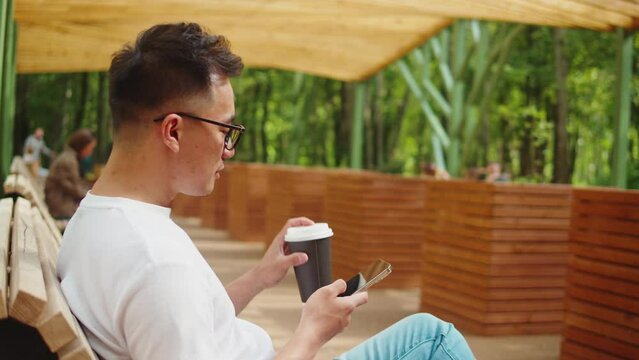 Asian man using phone outdoor close-up. Korean young guy freelancer working remotely in street. Remote work, business. Student communicating with friends online, chatting and drinking coffee.