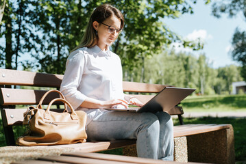 Education online, freelance outdoors. Cute female student wearing eyeglasses working on laptop while sitting on park bench. Young woman in casual clothes typing, browsing, studying information