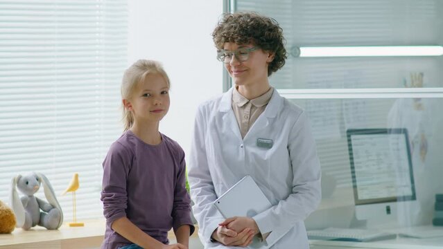 Portrait of pretty little girl and female doctor with digital tablet sitting together on medical couch in clinic and posing for camera