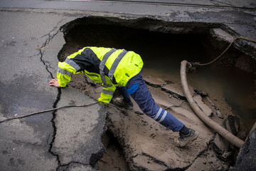 Road construction worker investigating Huge sinkhole on busy asphalt road surface on which cars...