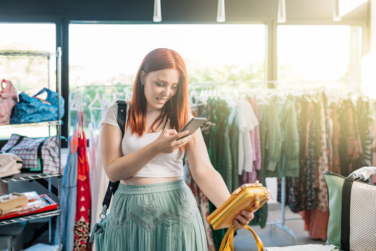 young red-haired woman happy shopping in a clothing shop, taking pictures with her smartphone of the clothes. girl spending time shopping sharing it on her social networks. concept of shopping.