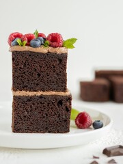 Two pieces of brownie cake on top of each other decorated chocolate cream cheese and raspberry, blueberry and mint leaves. White plate, white background, pieces of chocolate on the foreground