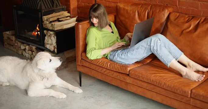Young woman works on laptop computer while sitting with her cute white dog on a couch at home. Remote work from home and friendship with pets