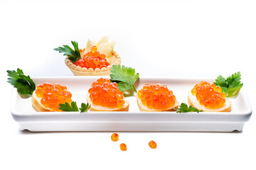 Luxurious red caviar scattered on white table. On small buns with butter lies caviar on sides decorated with parsley leaves. Buns lie on long white rectangular plate. At back is tartlet with caviar.