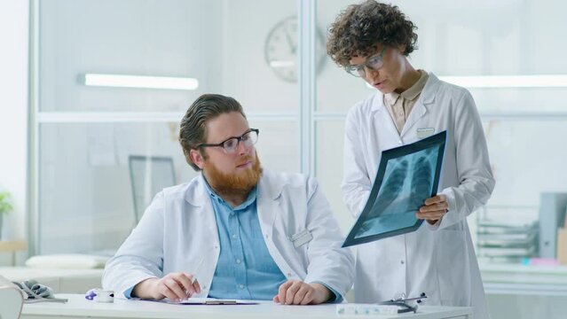 Male doctor in white coat working at desk in medical office and then discussing chest x-ray scan with female colleague