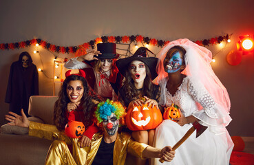 Happy adult friends in spooky costumes posing for funny group photo. Cheerful young people dressed as crazy witch, devil, vampire, dead bride and clown with axe having fun together at Halloween party - obrazy, fototapety, plakaty