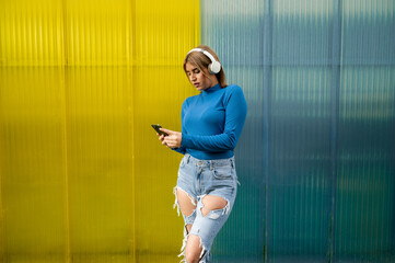 Young woman in casual clothes listening to music on the street with headphones while using the mobile phone on a blue and yellow background