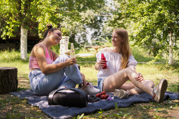 Two teenager girls have a picnic on a sunny day,  wearing summer clothes, sitting on a blanket, drinking a beverage surrounded by nature