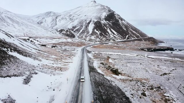 Aerial following flight behind driving white van on Djúpivogur route with massive snowy mountains at coastline