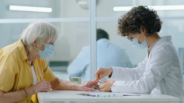 Female Doctor In Protective Face Mask Using Pulse Oximeter While Measuring Oxygen Level In Blood Of Senior Patient During Medical Checkup In Hospital In Times Of Pandemic