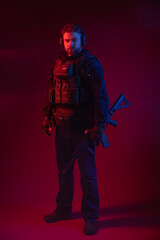 Fototapeta na wymiar soldier in full gear with weapons. a man in headphones, body armor, with a backpack and a belt. red background. colored, blue-red light