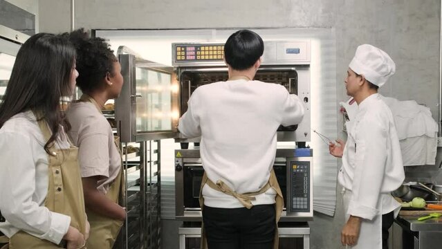 Senior Asian male chef of culinary learning course teaches and explains about electric oven to cooking class students for baking pastry dough for bread and bakery foods in stainless steel kitchen.