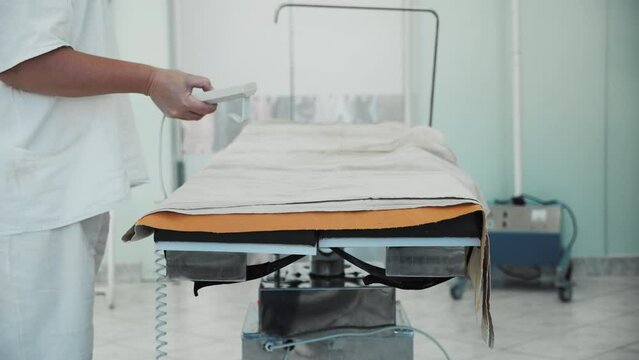 Close-up of scrub nurse pressing buttons on remote control, modern automatic motorized surgical operating table rotates and changes tilt angle, hi-tech equipment in hospital surgery operating room.