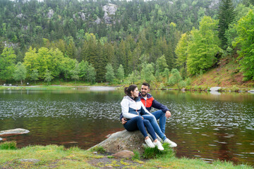 Couple in love looking into each other's eyes sitting on a rock with a lake and mountains with trees in the background.