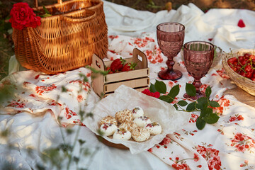 Feminine pink summer picnic with tablecloth, marshmallows, strawberries, cherry, peas and glasses of wine among pink roses in the park. Romantic evening, travel and appreciation of the moment.