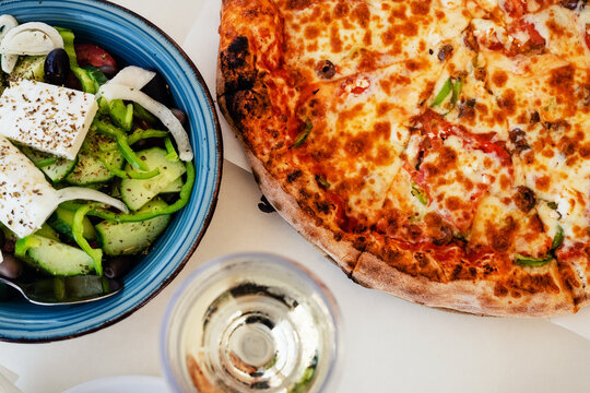 Traditional Greek Salad with feta cheese, Greek pizza and white wine glass in outdoor restaurant. Tourist Dinner in Agios Nikolaos in Crete, Greece. Summer vacation concept.