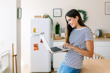 Smiling young caucasian woman typing on laptop while working from home - Freelancer entrepreneur female using computer in the kitchen - Small business and telecommuting concept