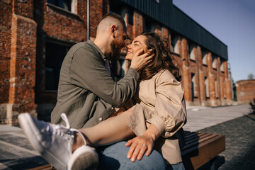 Portrait of sensual couple in casual clothes smiling and hugging together while sitting on bench in city street against brick wall.