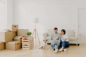 Affectionate married couple move in new home, relax on floor after unpacking things use laptop computer for searching home decoration ideas in internet their pet sits near, pose in purchased apartment