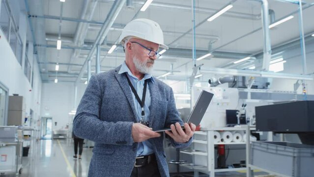 Portrait of a Bearded Middle Aged General Manager Walking in a Factory Facility, Wearing Casual Suit and a White Hard Hat. Heavy Industry Specialist Working on Laptop Computer.