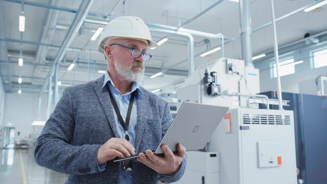 Portrait of a Bearded Middle Aged General Manager Standing in a Factory Facility, Wearing Casual Suit and a White Hard Hat. Heavy Industry Specialist Working on Laptop Computer.