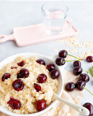 Oatmeal porridge with fresh black cherry in a white bowl with spoon and cup of water on white table. Healthy eating concept. Close-up. Selective focus. Blurred background.