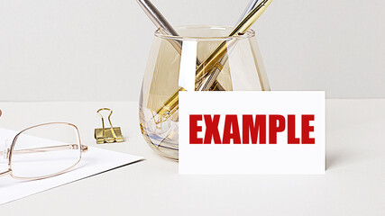 Work desk with gold glasses, pen, white card with the text EXAMPLE. Business concept. Home Office. Workplace close-up.