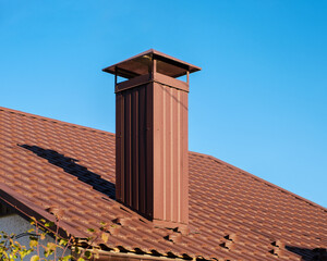 Close up shot of roof made of light brown metal tile, metal roofing shingles and chimney against blue sky.