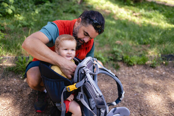 Fototapeta premium Young Caucasian father puts his little girl in a baby carrier. They are Going on a hiking adventure in a nearby forest. It's a lovely sunny summer day