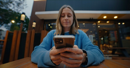 Attractive young woman with light hair wearing blue sweater, sitting outside in cafe, using smartphone, smiling, typing, texting, chatting with friends on social media, scrolling news on mobile phone