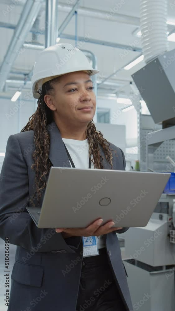 Poster Vertical Portrait of a Black Female Engineer in Hard Hat Walking and Using Laptop Computer at Electronic Manufacturing Factory. Technician Working on Daily Tasks and Research and Development Data.