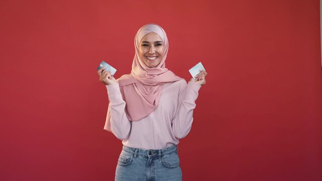 Banking service. Money transferring. Online shopping. Happy excited smiling woman in hijab holding credit card isolated on red studio background.