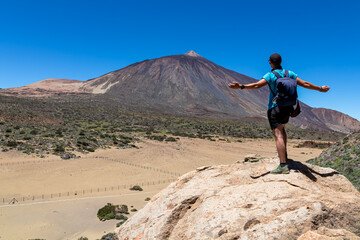 Fototapeta premium Man on rock with view on La Canada de los Guancheros dry desert plain and volcano Pico del Teide, Mount Teide National Park, Tenerife, Canary Islands, Spain, Europe. Hiking to Riscos de la Fortaleza