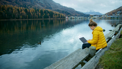 Fototapeta na wymiar Young woman in bright yellow jacket work on side of lake on her laptop. Female freelancer work remotely during vacation. Cinematic modern millennial work style concept