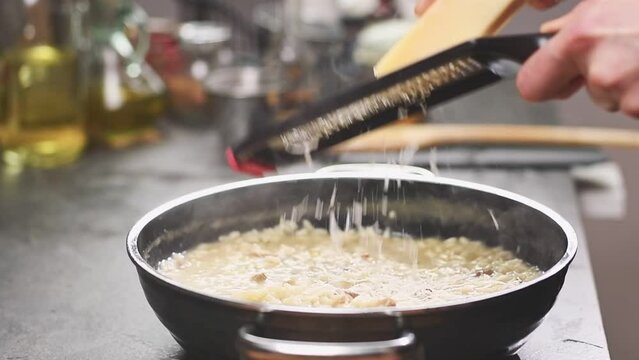 The cook grating parmesan cheese on risotto with mushrooms in the frying pan, slow motion.