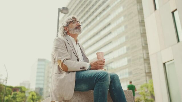 Mature businessman with beard in eyeglasses wearing gray jacket holding takeaway coffee. Middle aged manager enjoying morning coffee while sitting outside the office