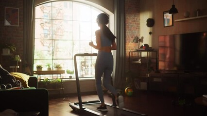 Beautiful Athletic Sports Woman Running on a Treadmill at Her Home Gym. Energetic Workout Female Training while Listening Podcast or Music in Headphones. Apartment with Window View. Side Back View